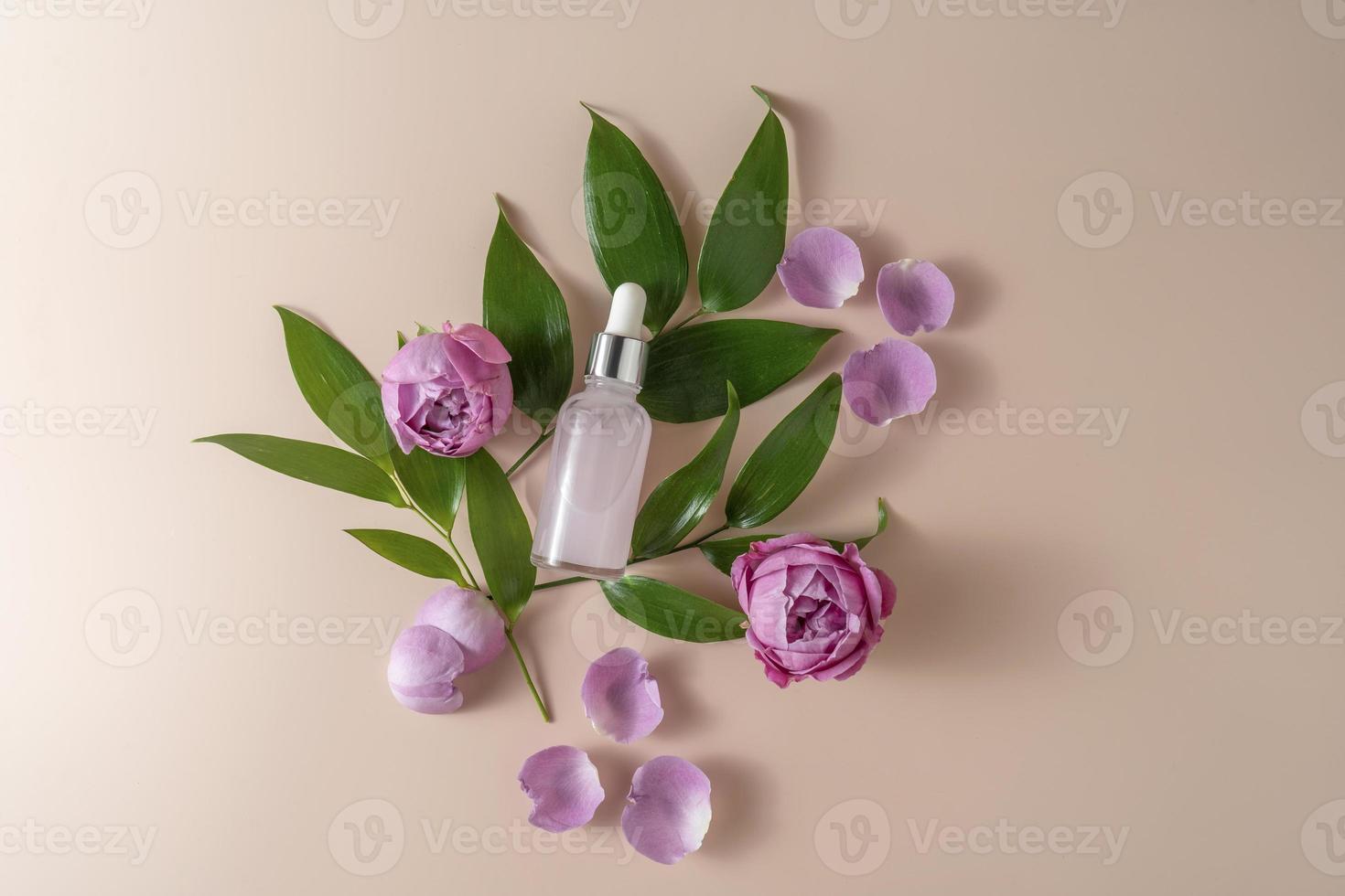 A face serum or essential oil in a pink dropper bottle lying on a beige background with rose petals and green leaves around it photo