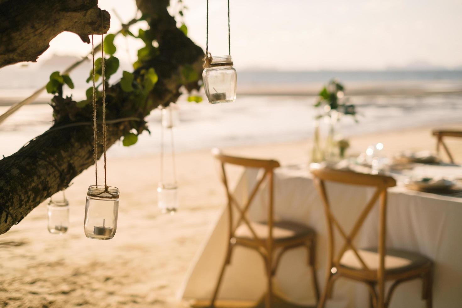 Cierra la vela del tarro que cuelga del árbol con una mesa larga para la cena de bodas en la playa de Tailandia por la noche. concepto de fiesta de bodas. decoración restaurante al aire libre en la playa. foto