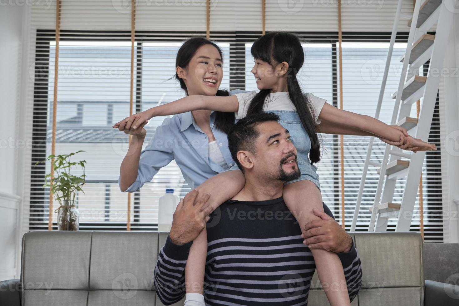 familia asiática tailandesa junta, papá juega y bromea con su hija y su madre cargando y sosteniendo a la niña sobre los hombros en la sala de estar de la casa, tiempos de ocio felices, fin de semana encantador, estilo de vida doméstico de bienestar. foto