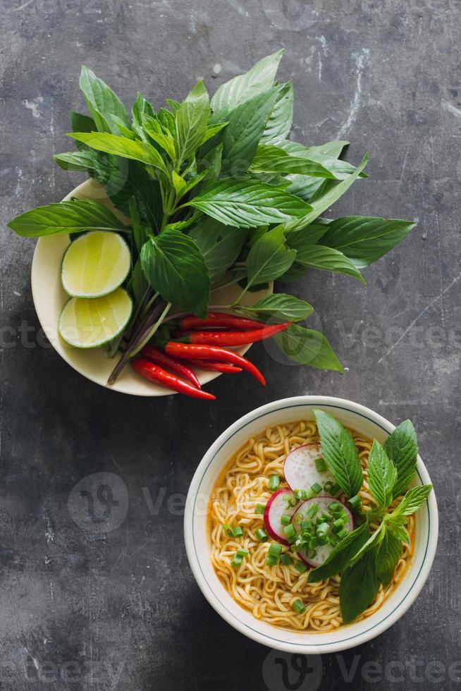 Instant noodles in bowl with fresh herbs, garnish of cilantro and Asian basil, lemon, lime on dark stone background photo