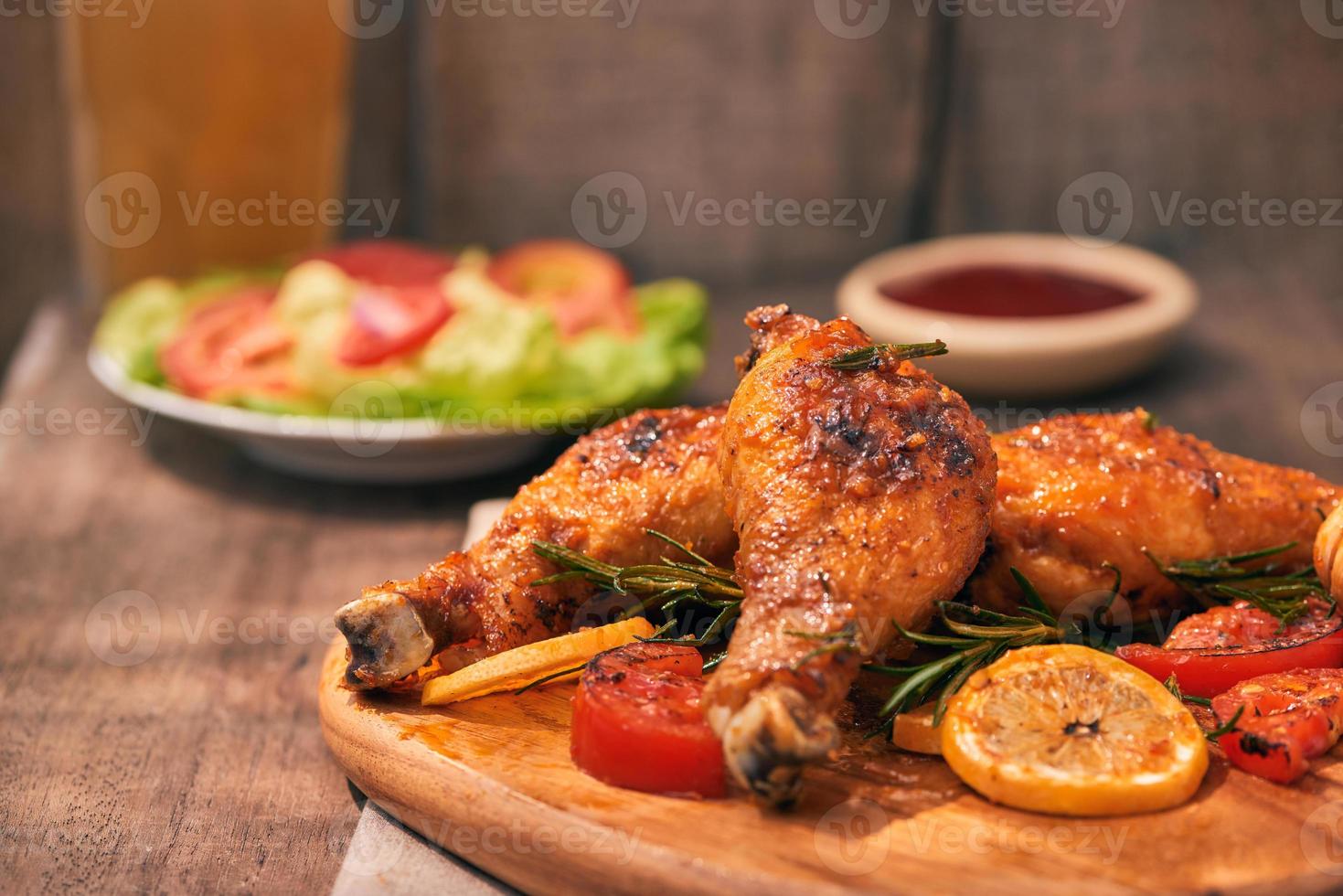 Grilled chicken legs roasted on the grill on wooden chopping board with tomato sauce in a bowl, fresh tomatoes and lettuce leaves, bitter pepper, glass mug of beer photo