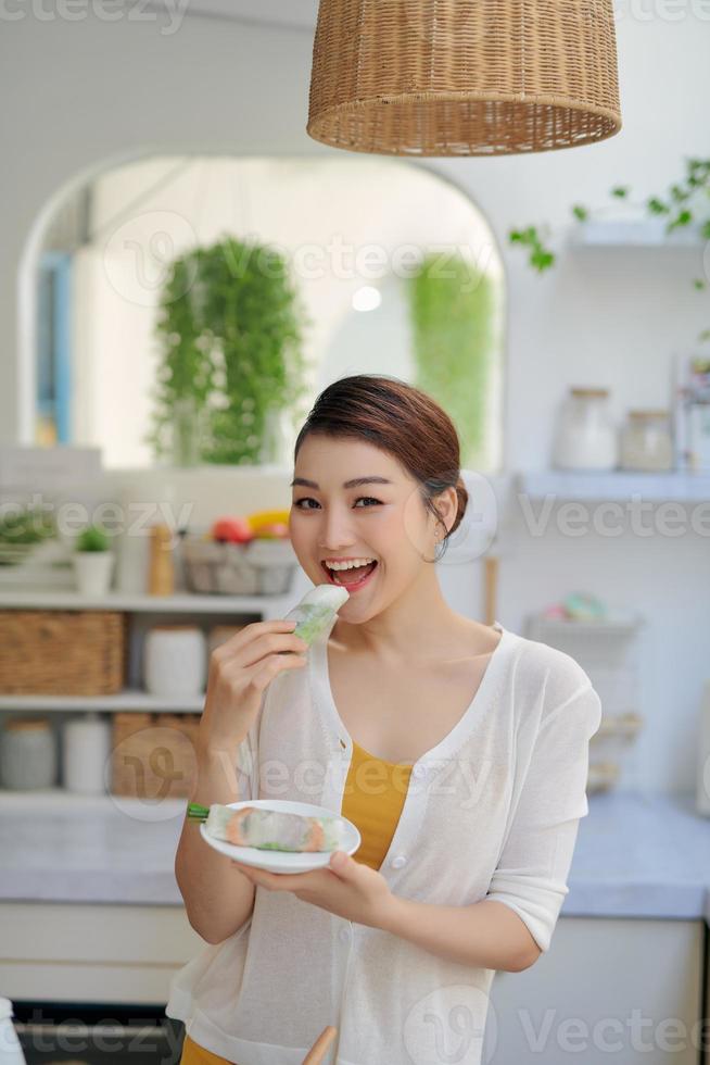 Asian woman eating spring rolls on kitchen background photo