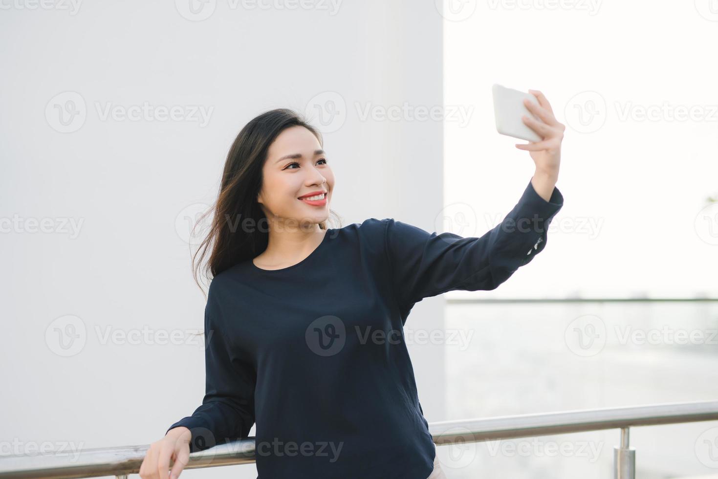 Relaxed and cheerful. Work and vacation. Outdoor portrait of happy young woman using smartphone, doing selfie photo and looking at you on terrace with beautiful city view.