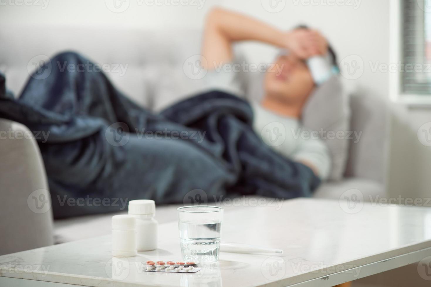 sick wasted man lying in sofa suffering cold and winter flu virus having medicine tablets in health care concept looking temperature on thermometer photo