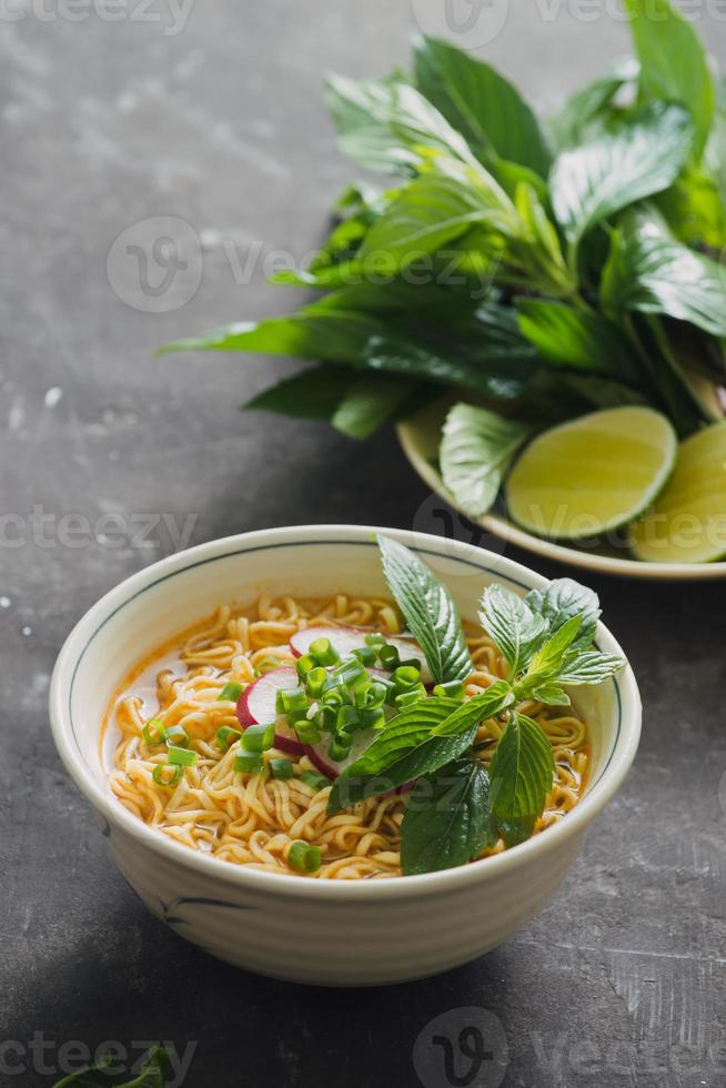 fideos instantáneos en tazón con hierbas frescas, guarnición de cilantro y albahaca asiática, limón, lima sobre fondo de piedra oscura foto