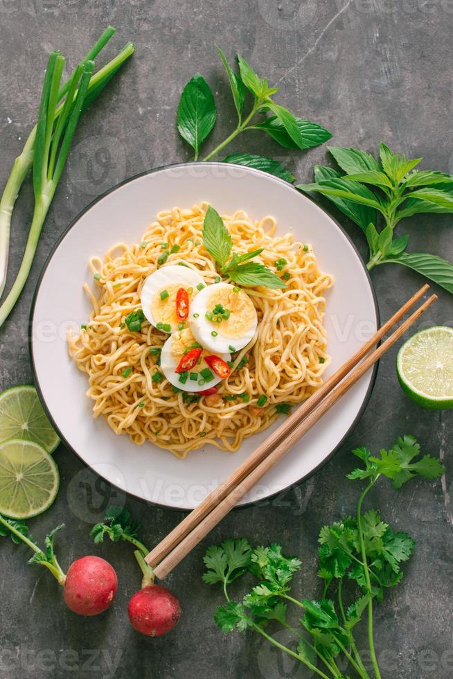 fideos instantáneos secos poner huevo con hierbas frescas, guarnición de cilantro y albahaca asiática, limón, lima sobre fondo de piedra oscura foto