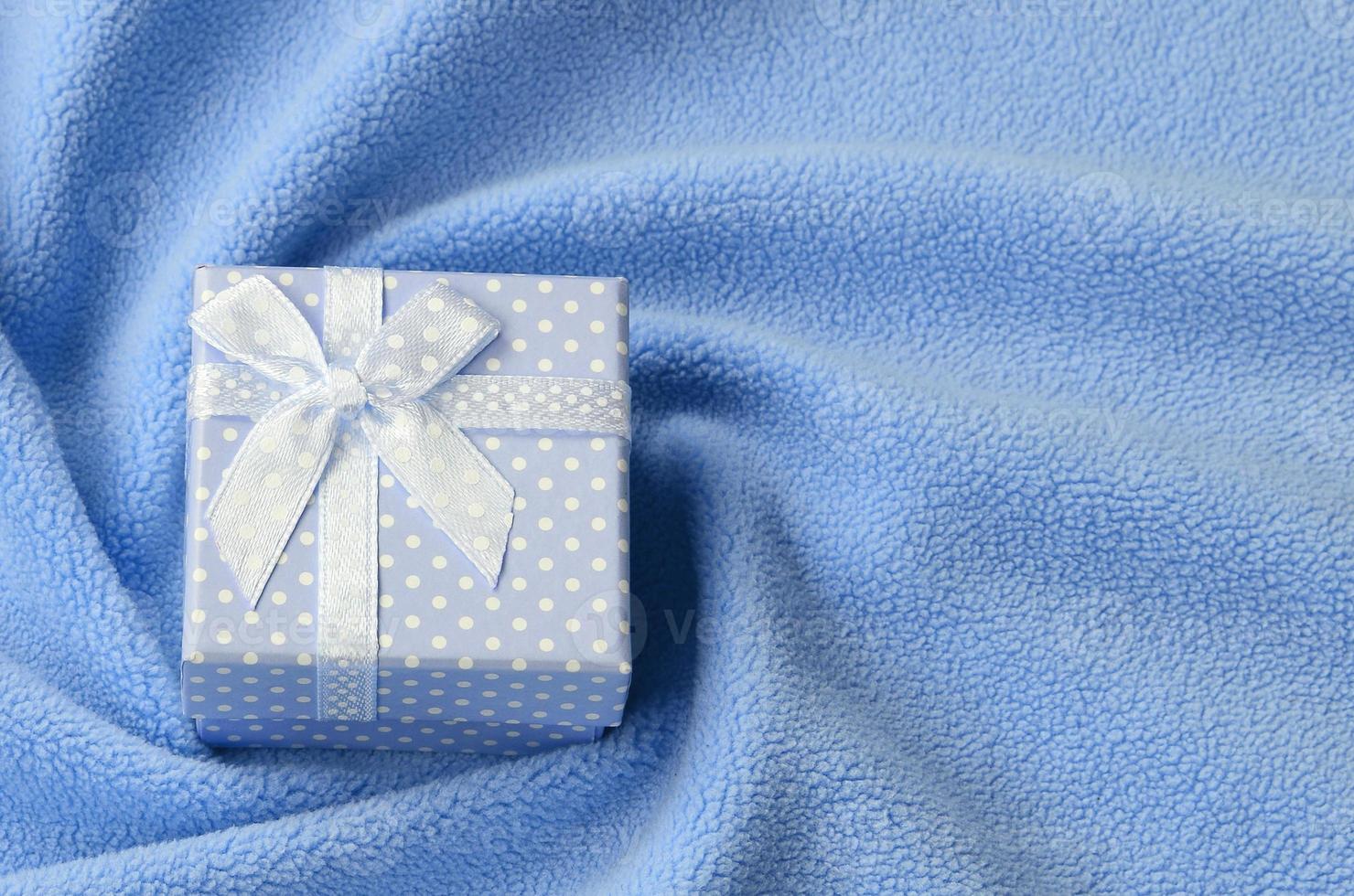 A small gift box in blue with a small bow lies on a blanket of soft and furry light blue fleece fabric with a lot of relief folds. Packing for a gift to your lovely girlfriend photo