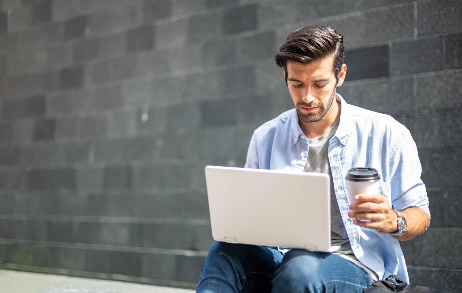 Young caucasian man holding cup of coffee and working on freelance project using portable computer at outer office. photo