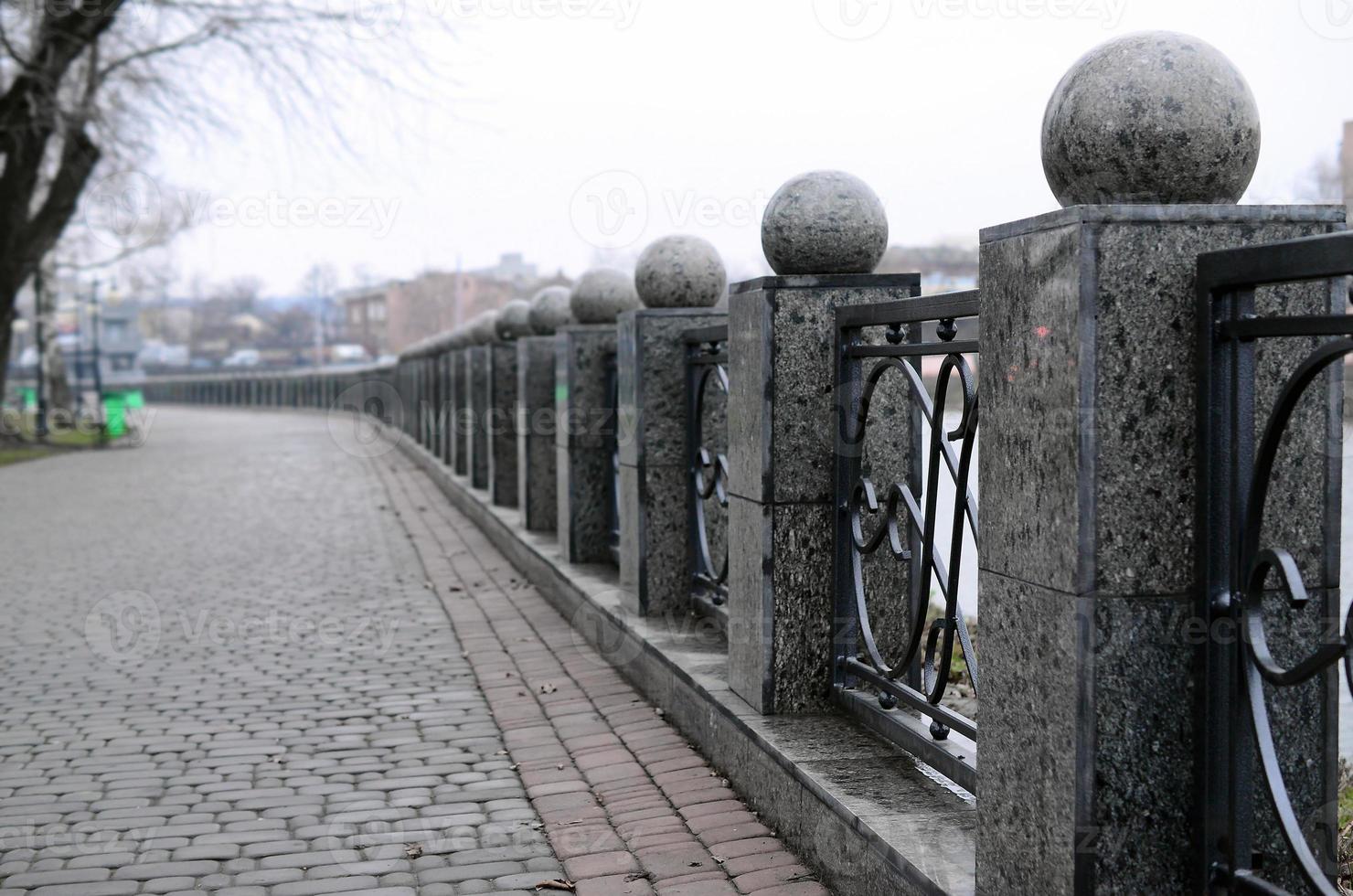 A beautiful granite fence with forged metal sections and decorative balls as decorations. The fence is built along the embankment of the street photo