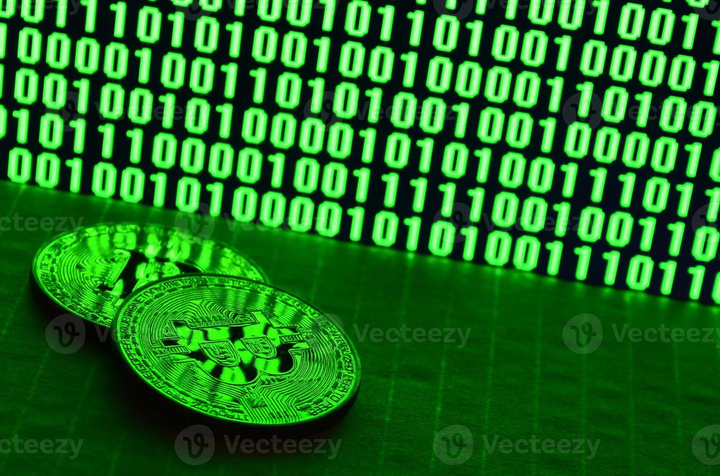 A pair of bitcoins lies on a cardboard surface on the background of a monitor depicting a binary code of bright green zeros and one units on a black background. Low key lighting photo