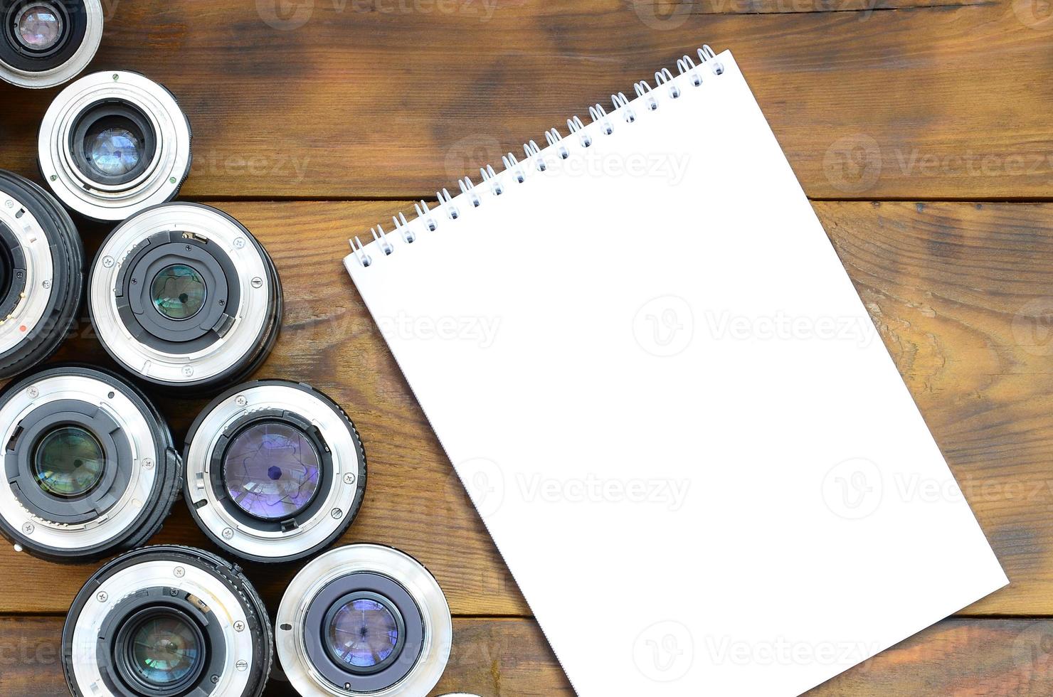 Several photographic lenses and white notebook lie on a brown wooden background. Space for text photo