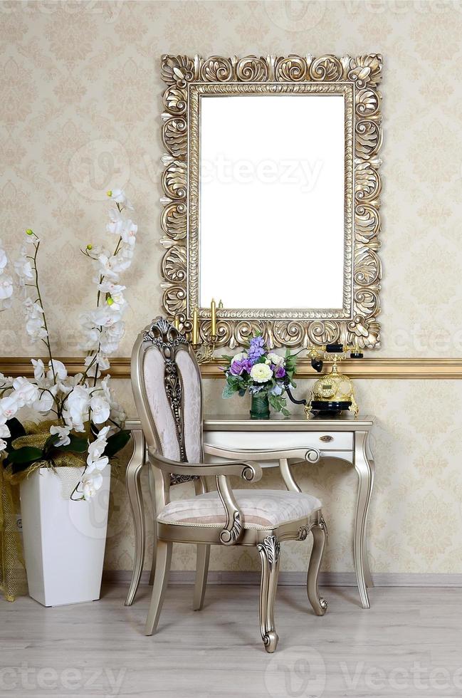 A fragment of a retro interior with a chair and table, on which is a telephone and a vase of flowers. Above the table hangs an empty picture frame for your photo