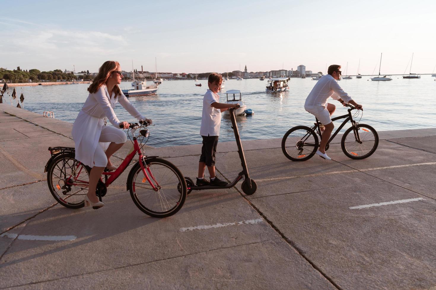 familia feliz disfrutando juntos de una hermosa mañana junto al mar, padres montando en bicicleta y su hijo montando una scooter eléctrica. enfoque selectivo foto