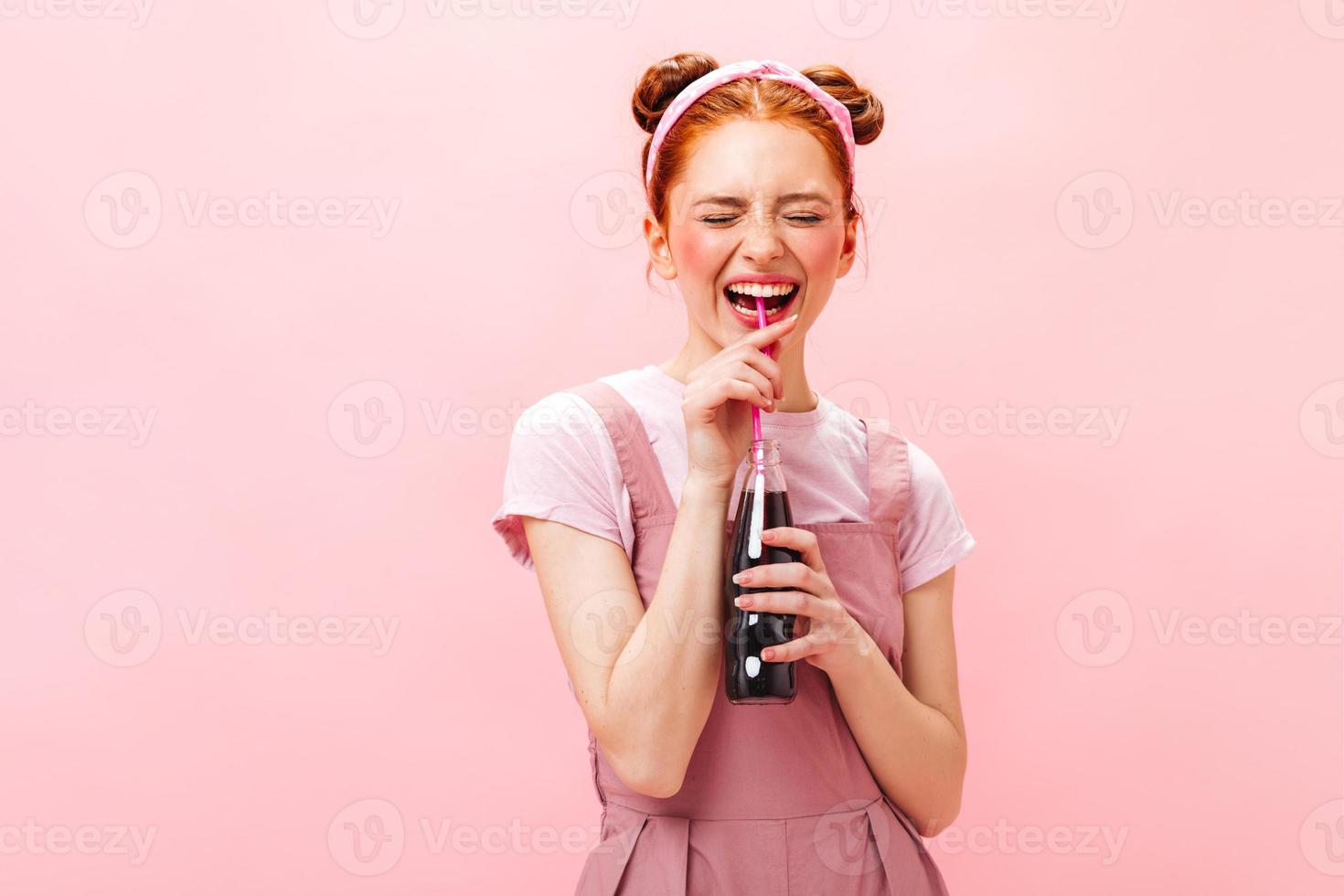 mujer pelirroja con vestido rosa y top blanco bebe refrescos a través de paja y posa sorprendida en isolat foto