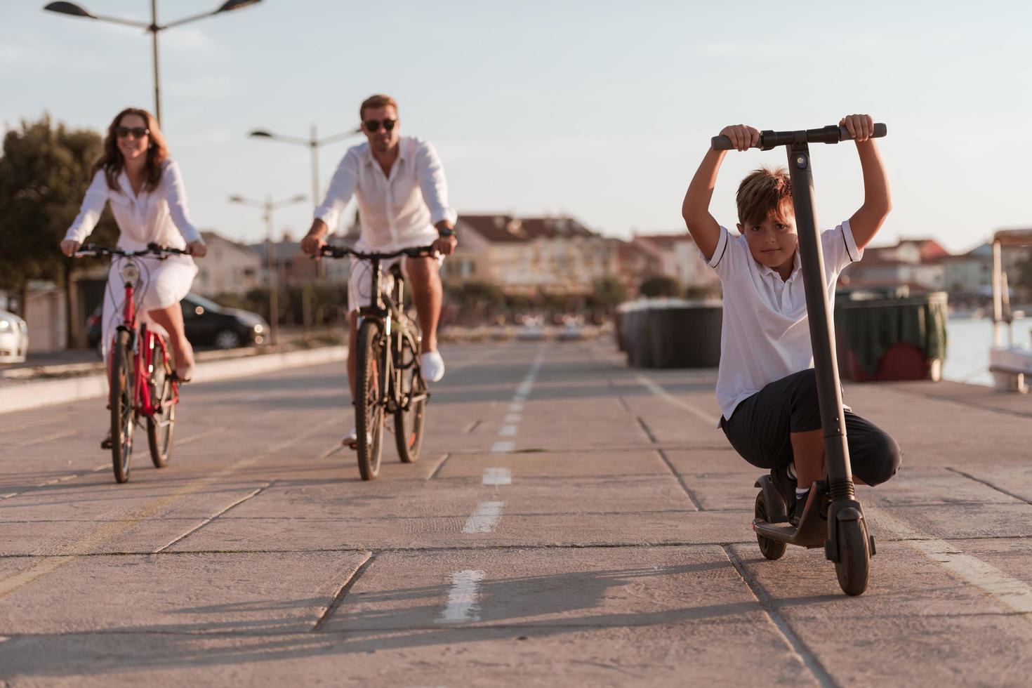 Happy family enjoying a beautiful morning by the sea together, parents riding a bike and their son riding an electric scooter. Selective focus photo
