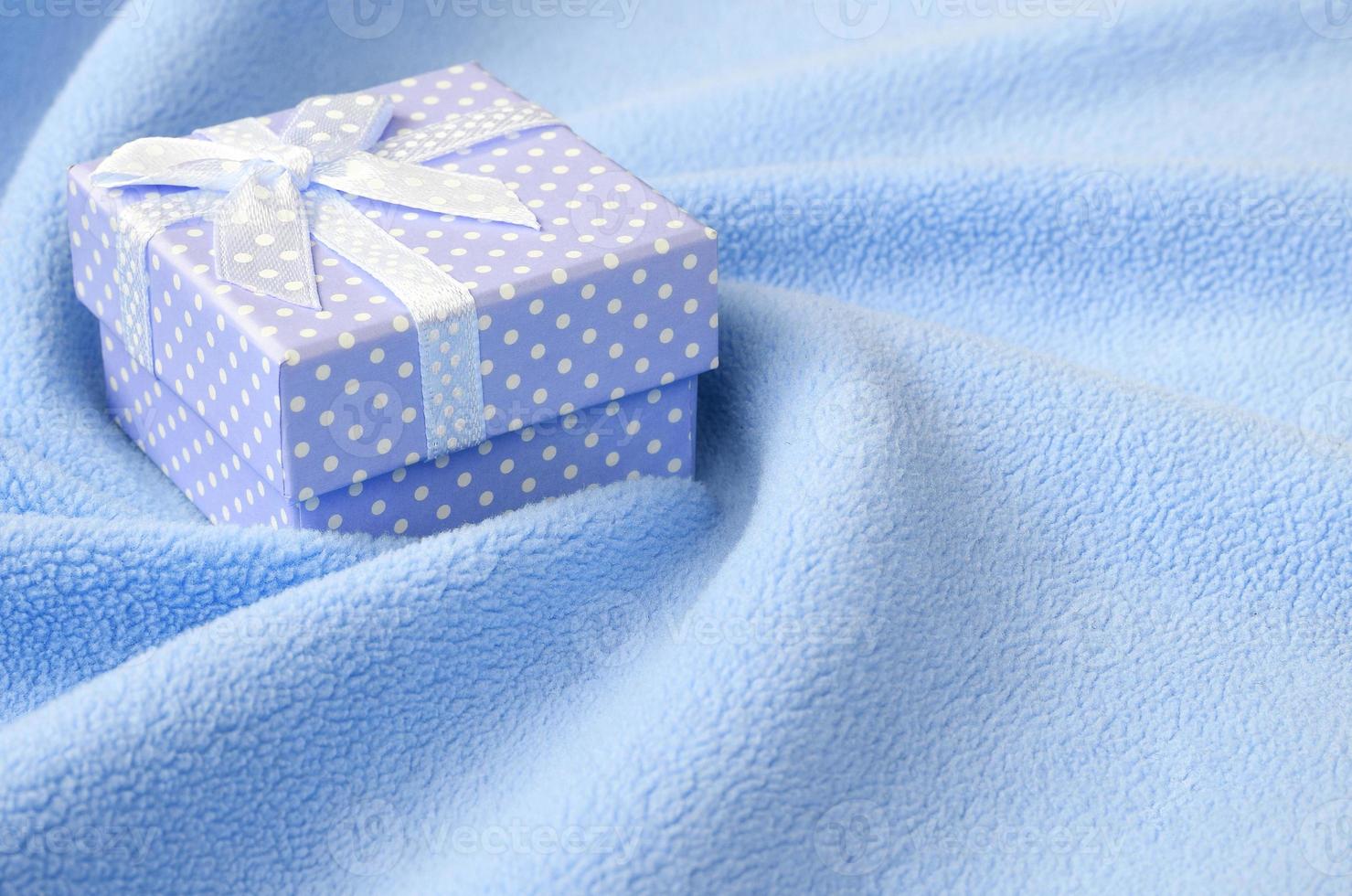 A small gift box in blue with a small bow lies on a blanket of soft and furry light blue fleece fabric with a lot of relief folds. Packing for a gift to your lovely girlfriend photo