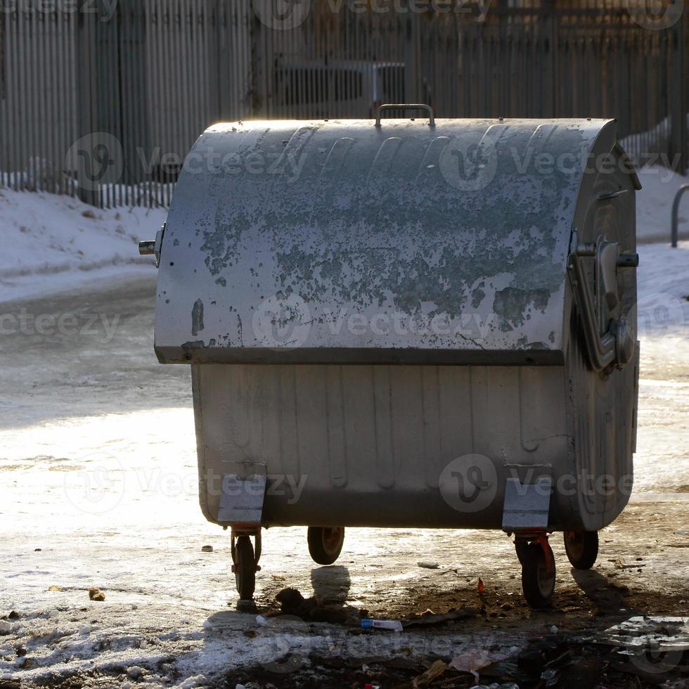 A silver garbage container stands near residential buildings in winter photo