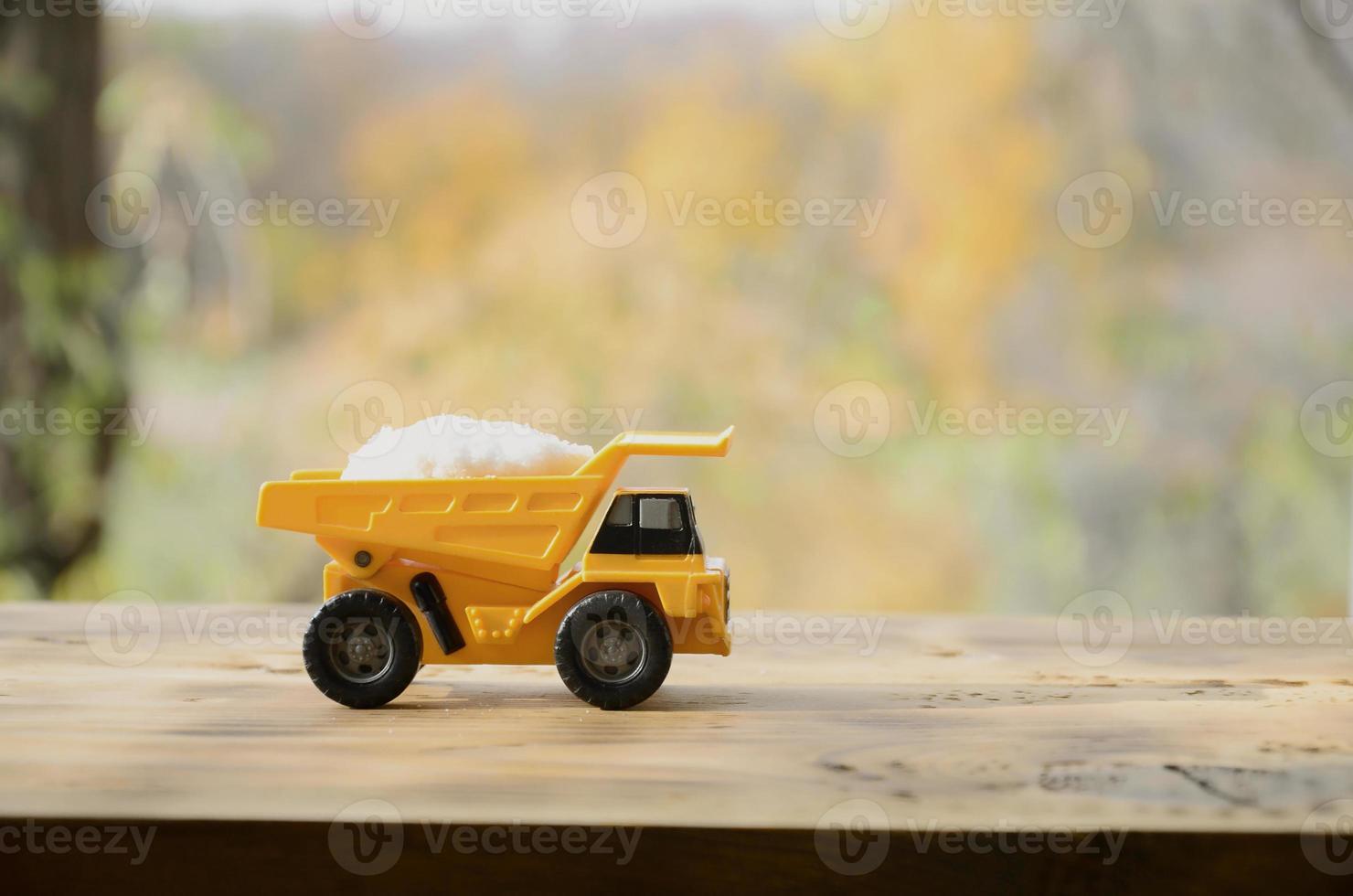 A small yellow toy truck is loaded with a stone of white salt. A car on a wooden surface against a background of autumn forest. Extraction and transportation of salt photo