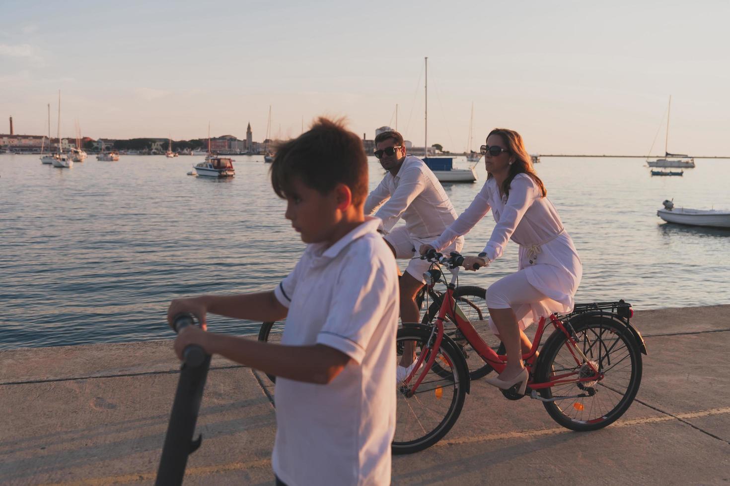 Happy family enjoying a beautiful morning by the sea together, parents riding a bike and their son riding an electric scooter. Selective focus photo