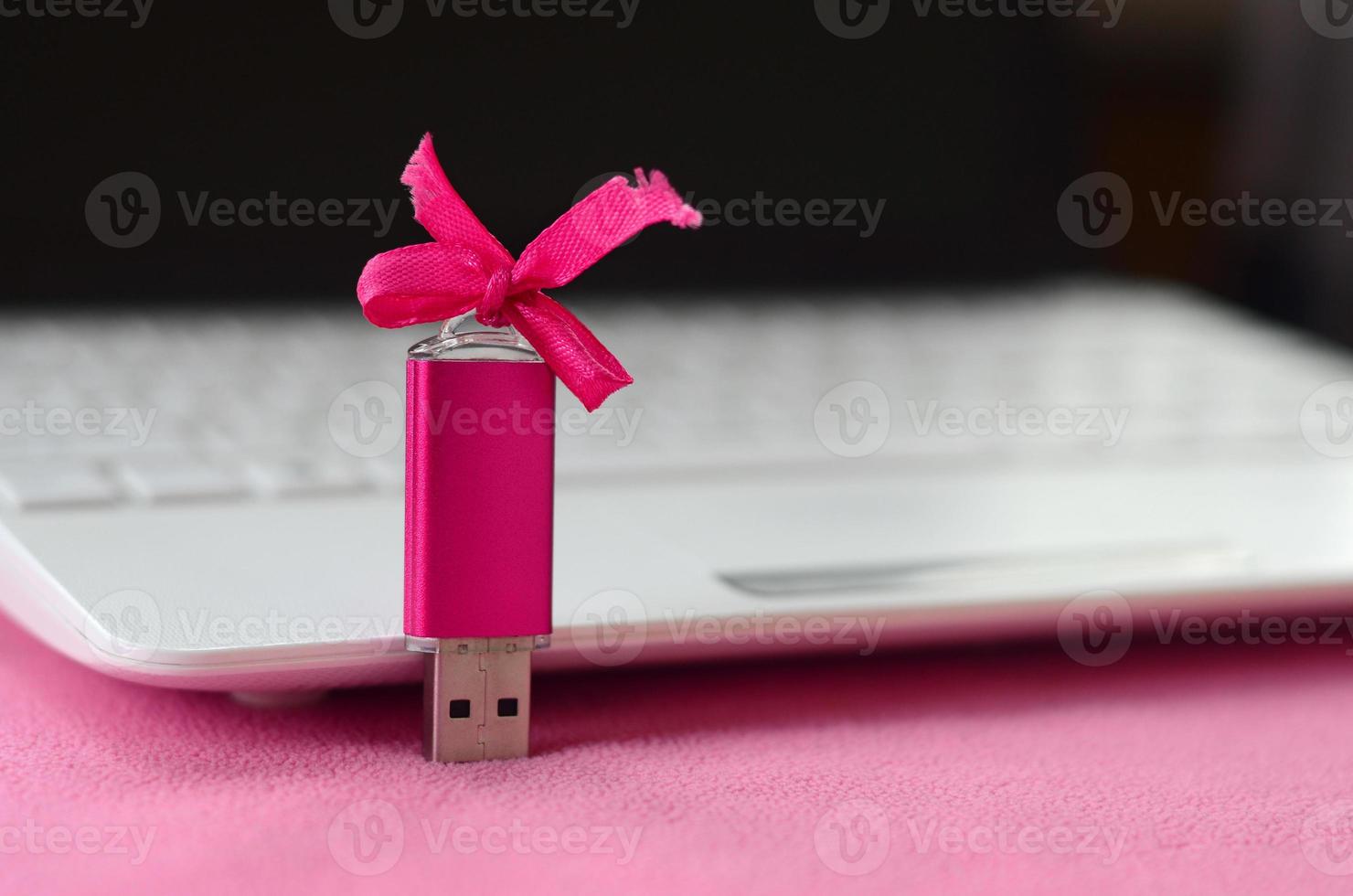 Brilliant pink usb flash memory card with a pink bow lies on a blanket of soft and furry light pink fleece fabric beside to a white laptop. Classic female gift design for a memory card photo