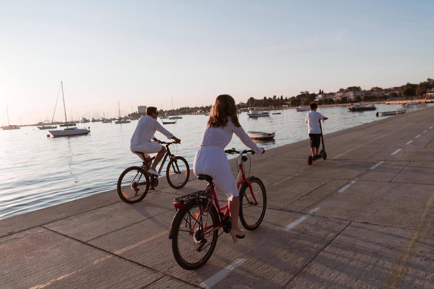 familia feliz disfrutando juntos de una hermosa mañana junto al mar, padres montando en bicicleta y su hijo montando una scooter eléctrica. enfoque selectivo foto