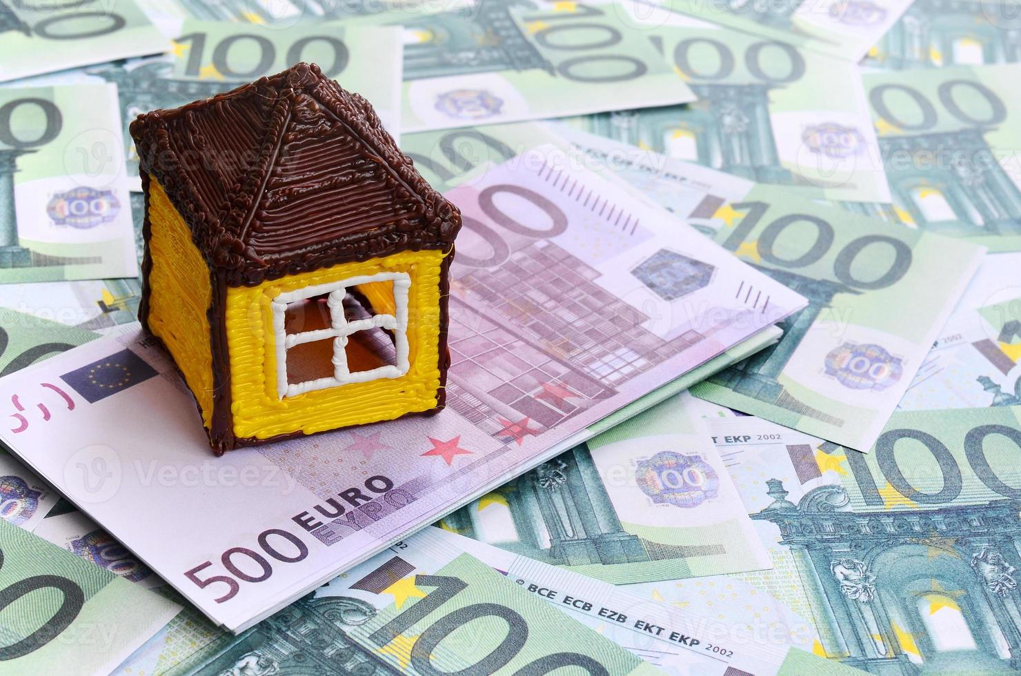 Small toy house is lies on a set of green monetary denominations of 100 euros. A lot of money forms an infinite heap photo