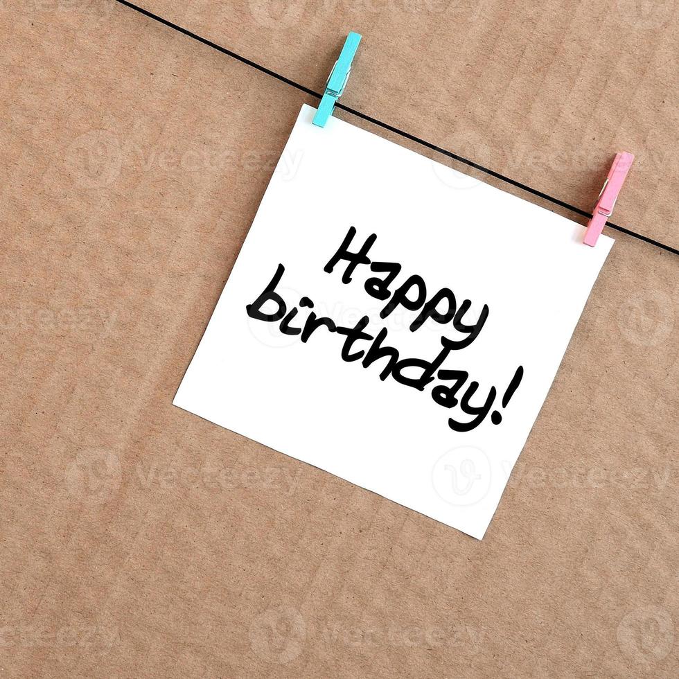 Happy birthday Note is written on a white sticker that hangs with a clothespin on a rope on a background of brown cardboard photo