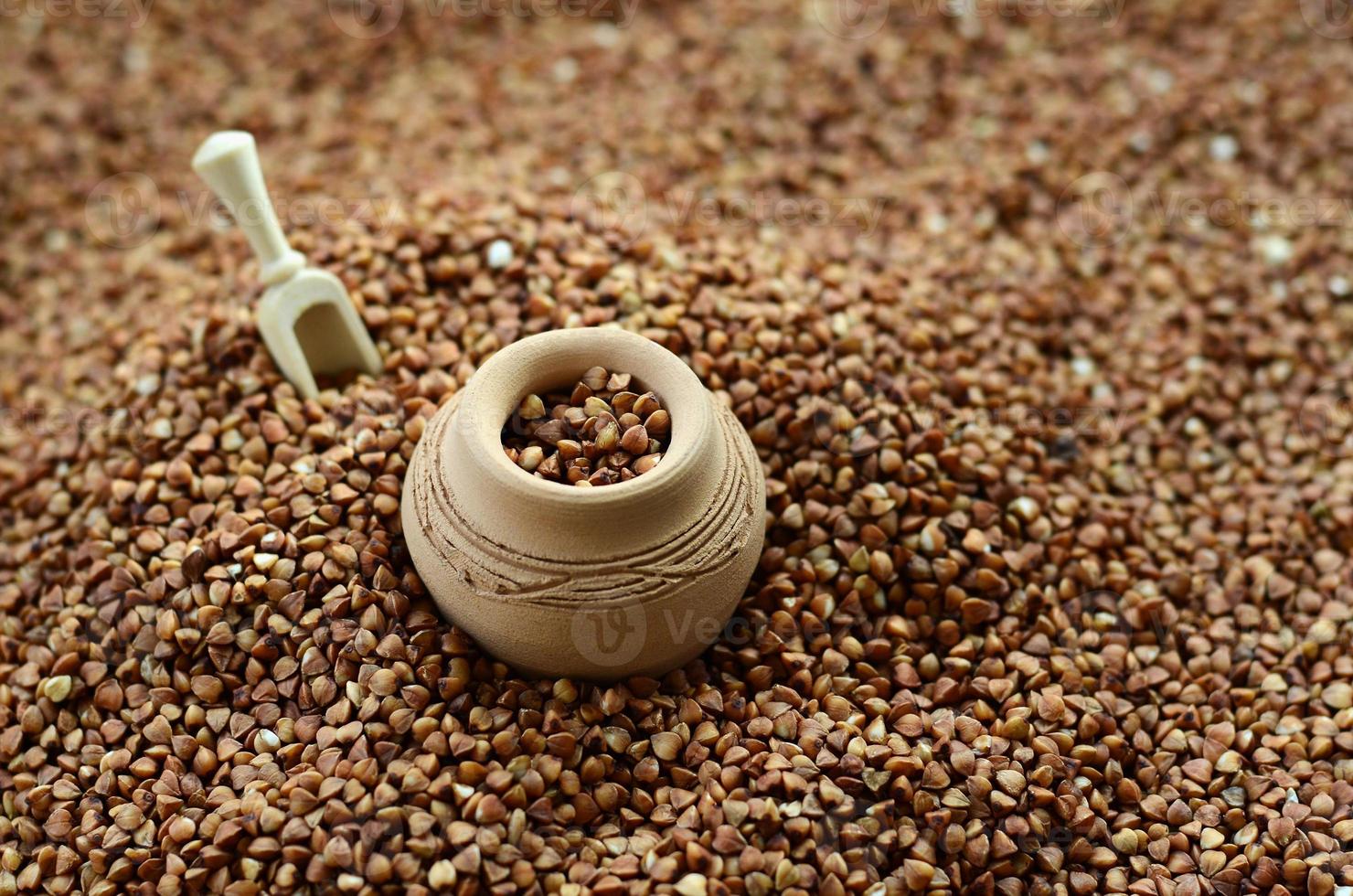 Background image of a large pile of buckwheat, in the middle of which lies a small jug and a wooden spatula for cereals photo