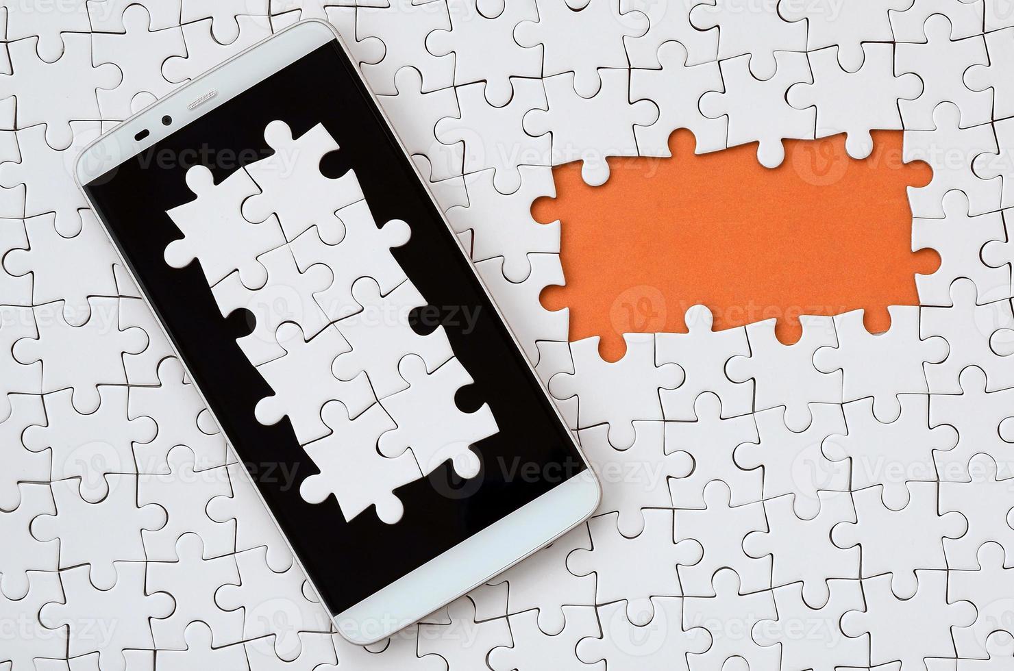 A modern big smartphone with several puzzle elements on the touch screen lies on a white jigsaw puzzle in an assembled state with missing elements photo