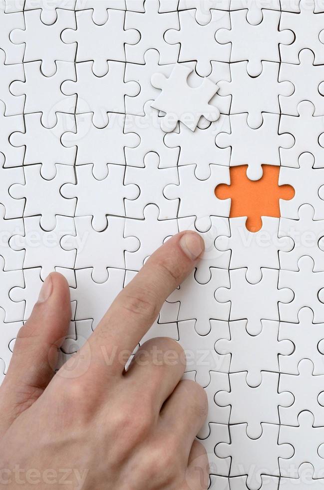 la textura de un rompecabezas blanco en estado ensamblado con un elemento faltante, formando un espacio naranja, señalado por el dedo de la mano masculina foto