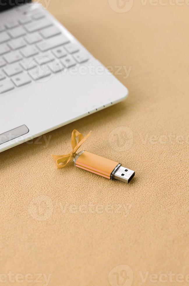 Orange usb flash memory card with a bow lies on a blanket of soft and furry light orange fleece fabric beside to a white laptop. Classic female gift design for a memory card photo