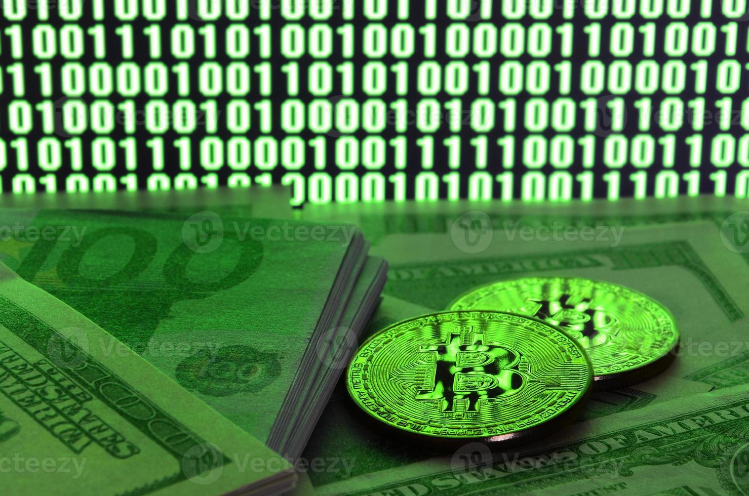 Two bitcoins lies on a pile of dollar bills on the background of a monitor depicting a binary code of bright green zeros and one units on a black background. Low key lighting photo