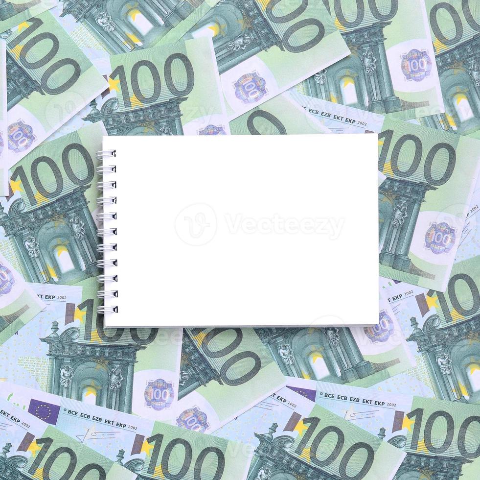 cuaderno blanco con páginas limpias sobre un conjunto de denominaciones monetarias verdes de 100 euros. mucho dinero forma un montón infinito foto