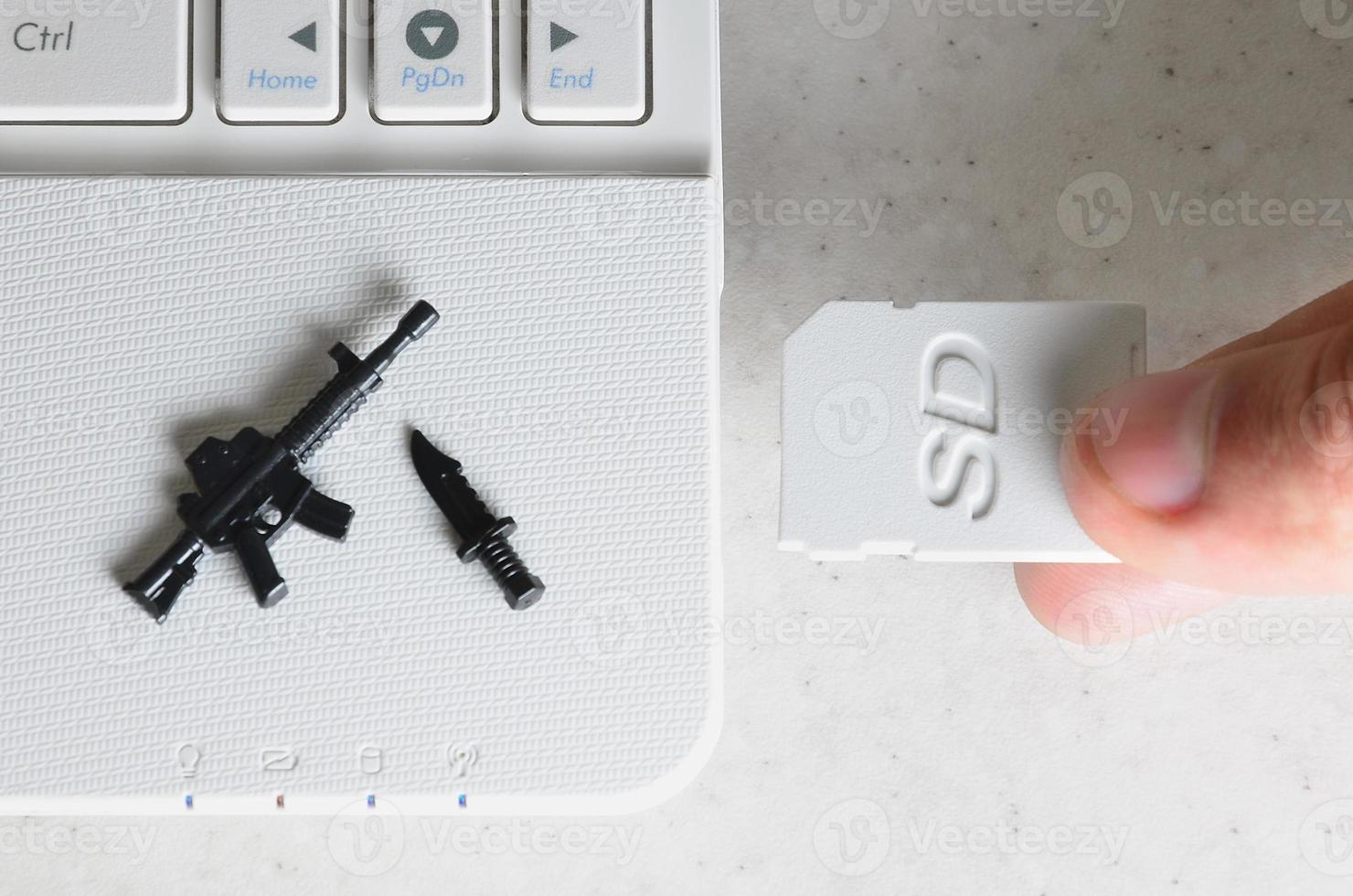A hand inserts a compact SD card into the slot of a netbook, on which lie a miniature knife and gun. The concept of information warfare, journalistic activity, dissemination of provocative information photo