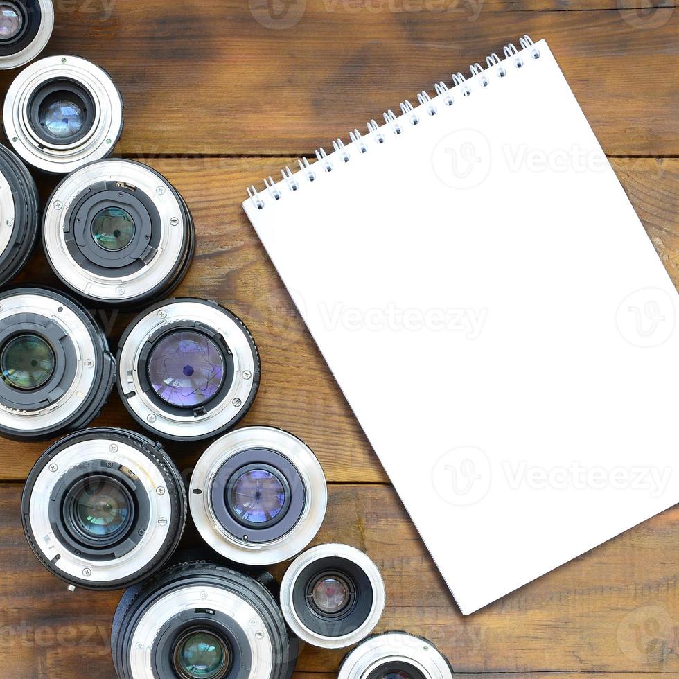 Several photographic lenses and white notebook lie on a brown wooden background. Space for text photo