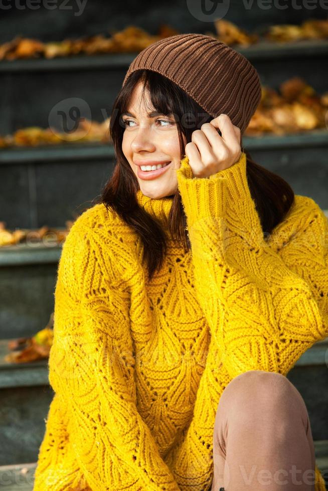 Beautiful European brunette dressed in a yellow woolen sweater and hat outside. The beautiful girl who wears thick stylish autumn clothes in cool weather. photo