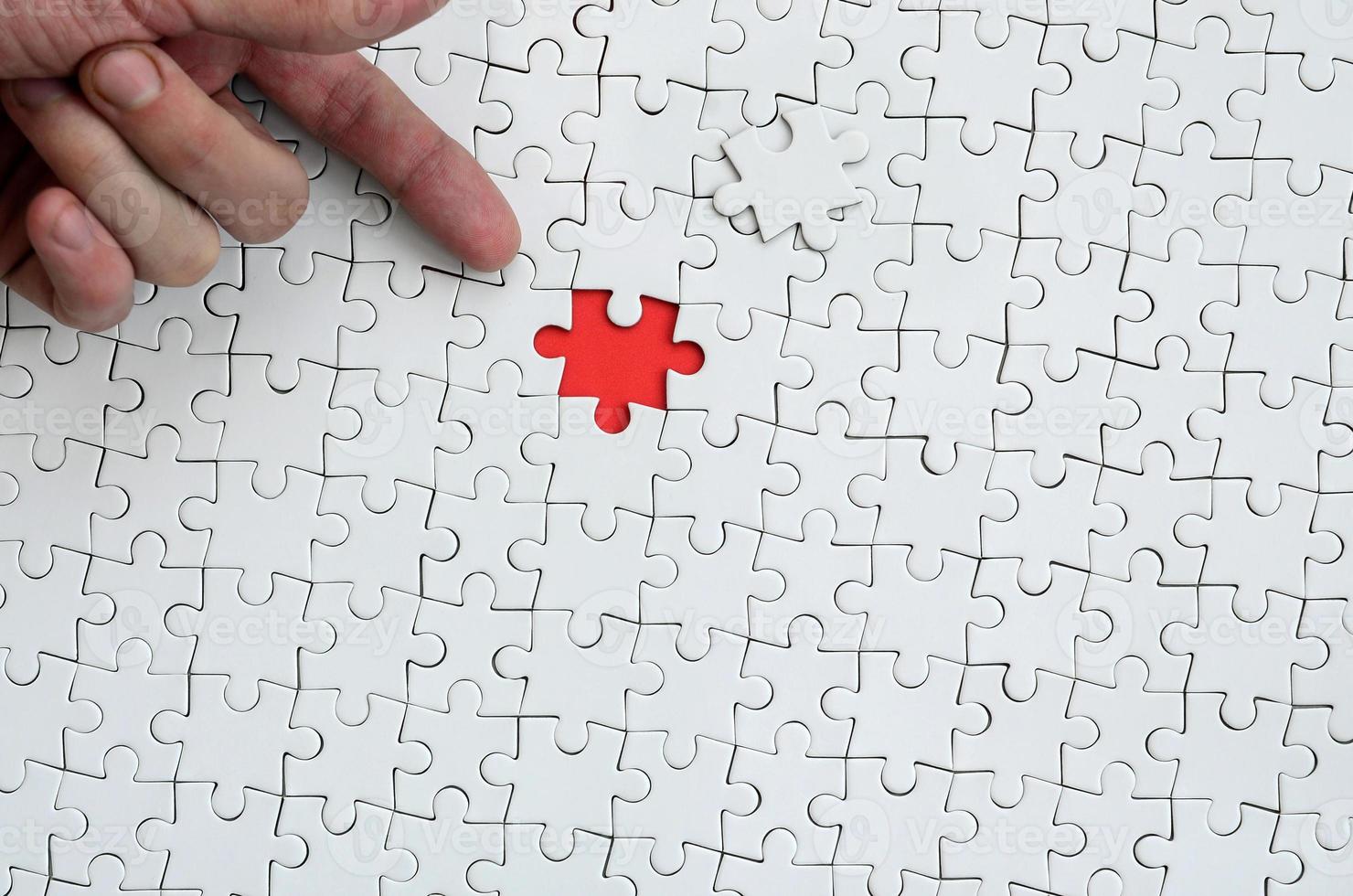 The texture of a white jigsaw puzzle in the assembled state with one missing element, forming a red space, pointed to by the finger of the male hand photo