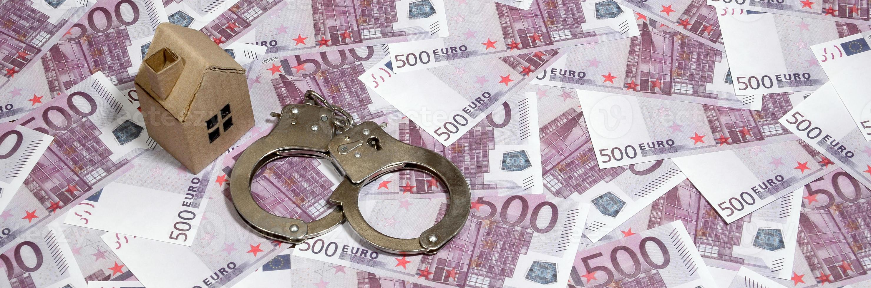 Scam with real estate. Cheating when buying or renting a house. Punishment for deception. A toy house with handcuffs on a many 500 purple euro banknotes photo