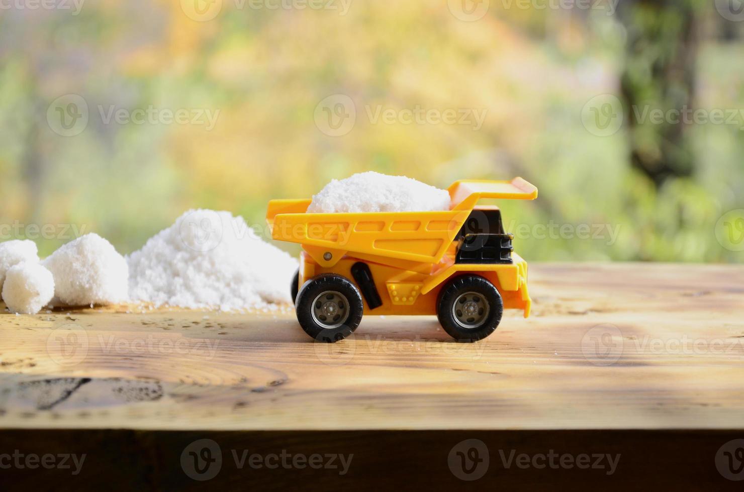A small yellow toy truck is loaded with a stone of white salt next to a pile of salt. A car on a wooden surface against a background of autumn forest. Extraction and transportation of salt photo