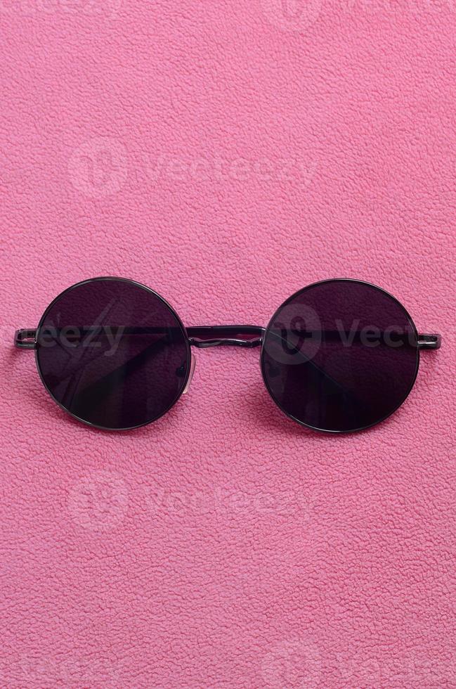 Stylish black sunglasses with round glasses lies on a blanket made of soft and fluffy light pink fleece fabric. Fashionable background picture in female colors photo