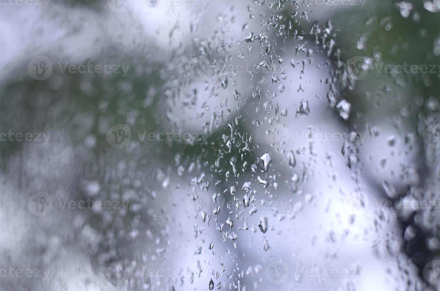 A photo of rain drops on the window glass with a blurred view of the blossoming green trees. Abstract image showing cloudy and rainy weather conditions