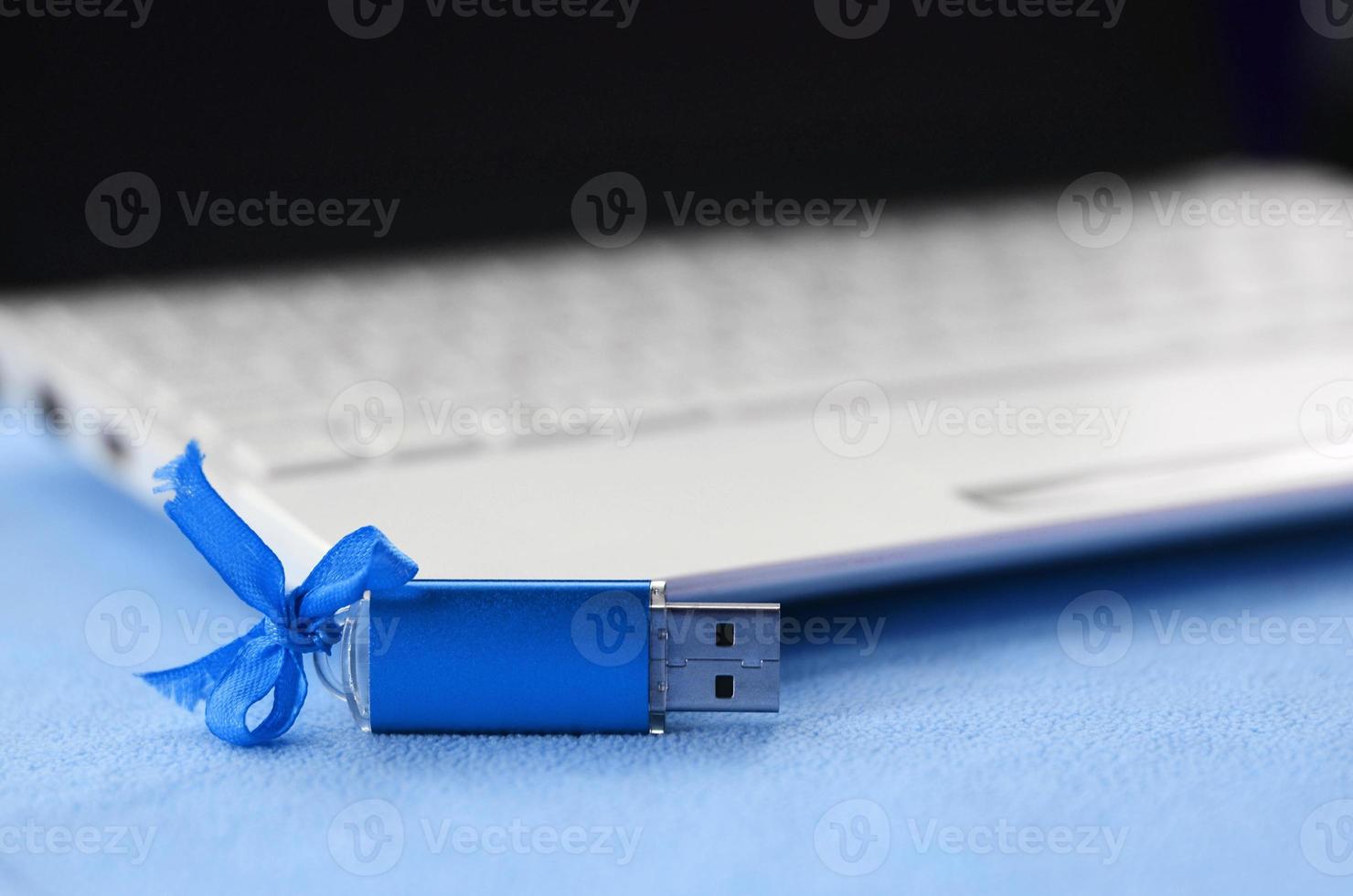 Brilliant blue usb flash memory card with a blue bow lies on a blanket of soft and furry light blue fleece fabric beside to a white laptop. Classic female gift design for a memory card photo