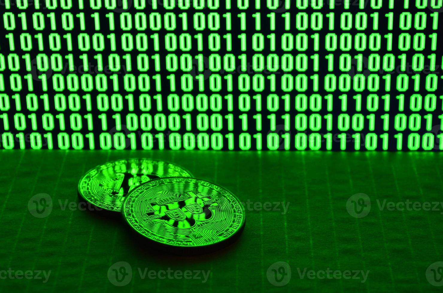 A pair of bitcoins lies on a cardboard surface on the background of a monitor depicting a binary code of bright green zeros and one units on a black background. Low key lighting photo