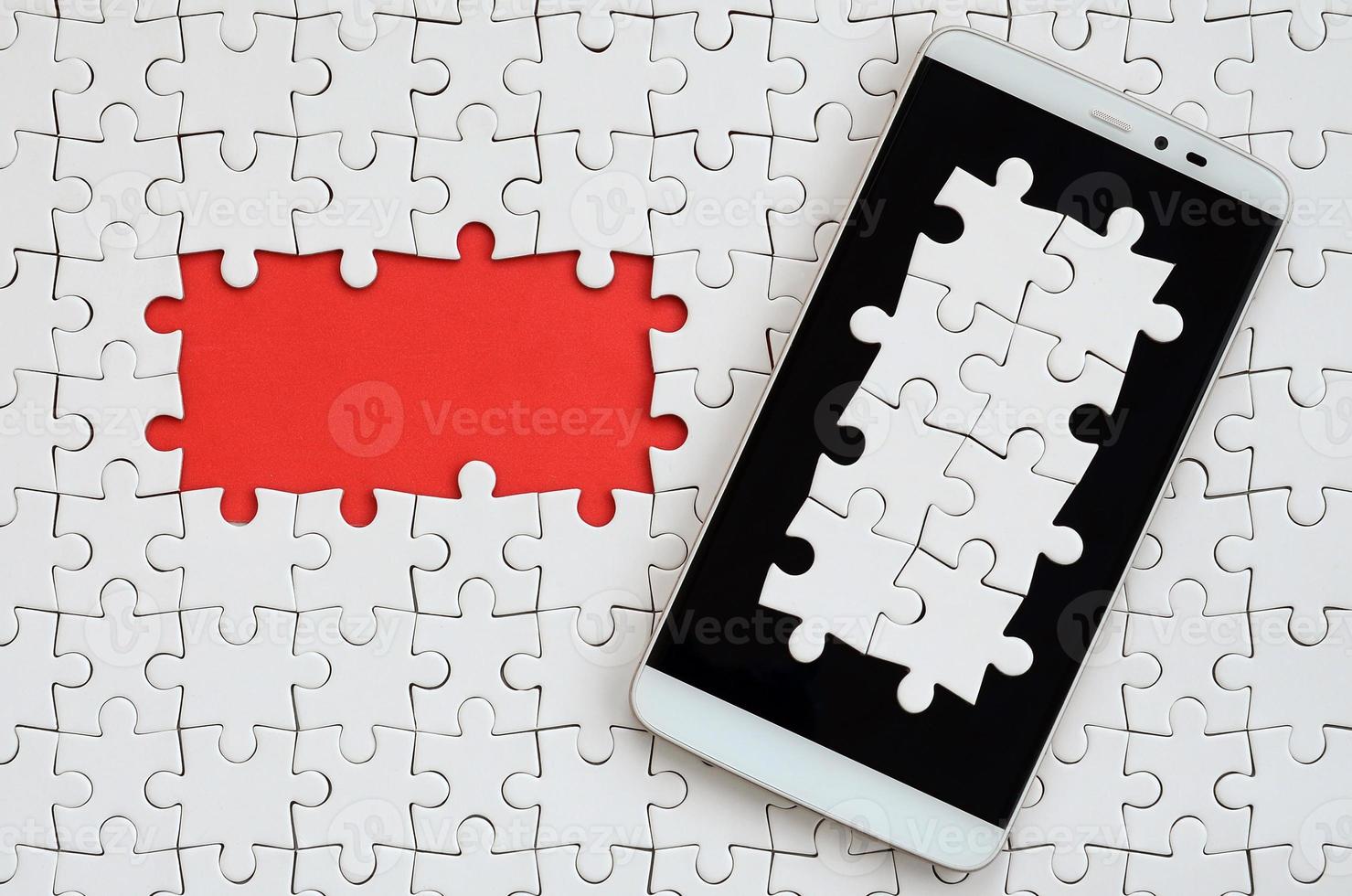 A modern big smartphone with several puzzle elements on the touch screen lies on a white jigsaw puzzle in an assembled state with missing elements photo