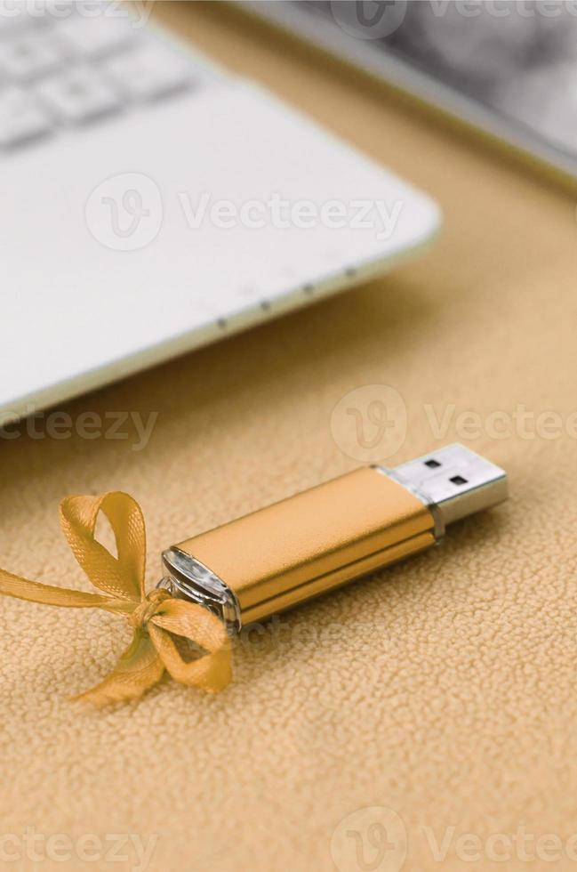 Orange usb flash memory card with a blue bow lies on a blanket of soft and furry light orange fleece fabric beside to a white laptop and smartphone. Classic female gift design for a memory card photo