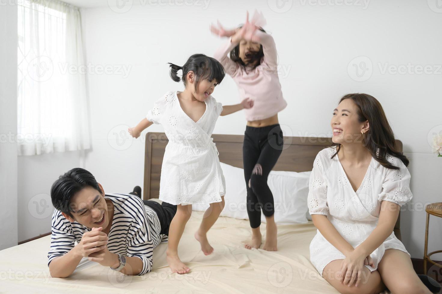 padres asiáticos con dos hijas pequeñas saltando en la cama en el dormitorio, concepto de familia feliz foto