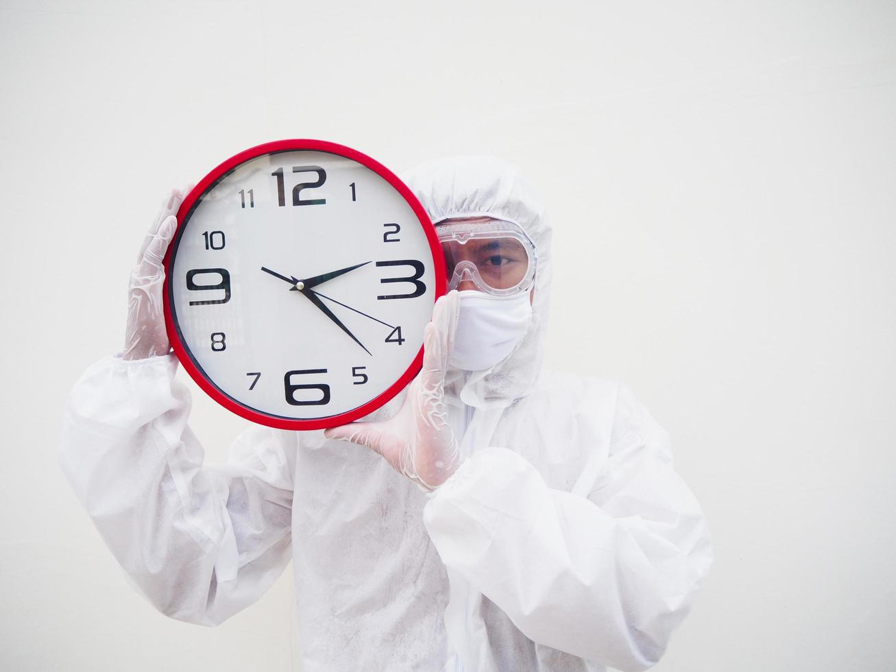 retrato de médico o científico con uniforme de suite ppe sosteniendo un despertador rojo y mirando la cámara con varios gestos. covid-19 concepto aislado fondo blanco foto