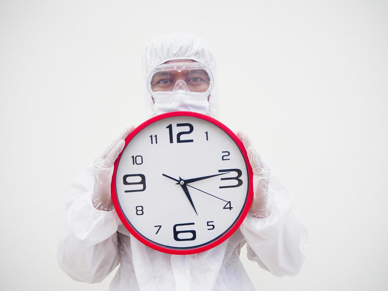 retrato de médico o científico con uniforme de suite ppe sosteniendo un despertador rojo y mirando la cámara con varios gestos. covid-19 concepto aislado fondo blanco foto