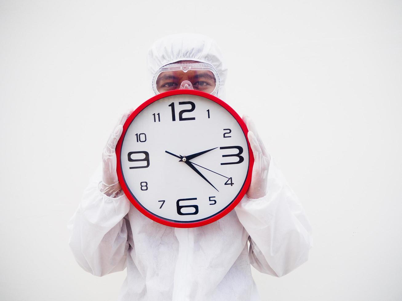 retrato de médico o científico con uniforme de suite ppe sosteniendo un despertador rojo y mirando la cámara con varios gestos. covid-19 concepto aislado fondo blanco foto