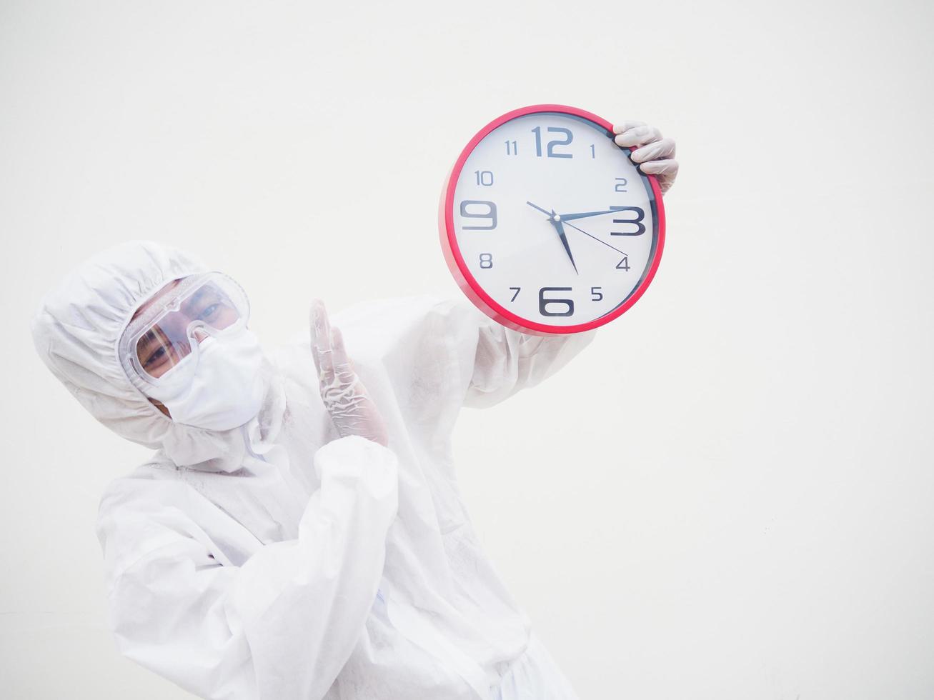 retrato de médico o científico con uniforme de suite ppe sosteniendo un despertador rojo y mirando la cámara con varios gestos. covid-19 concepto aislado fondo blanco foto